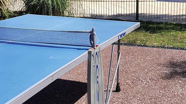 Una mesa de ping pong portátil sobre un terreno de grava, con una piscina y palmeras al fondo.