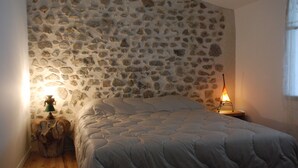 A bedroom with a stone wall, a bed with a quilt, a wooden nightstand with a lamp, and a window with curtains. - Charming and comfortable loft-style gîte*** near Vichy for 4 to 5 people (Châteldon)