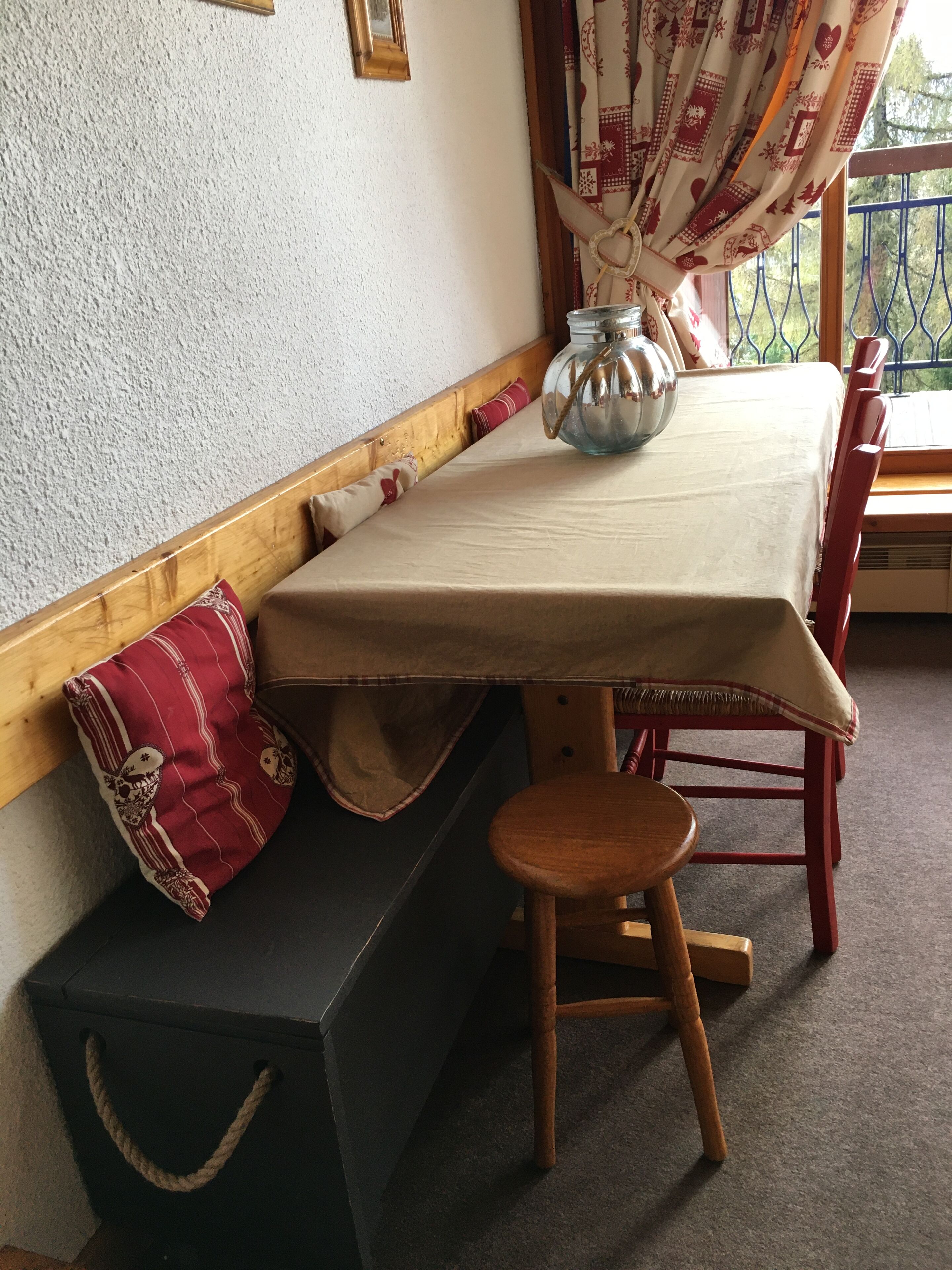 Een hoekkamer met een houten bankje, een klein krukje, een tafel met tafelkleed en een decoratieve vaas.