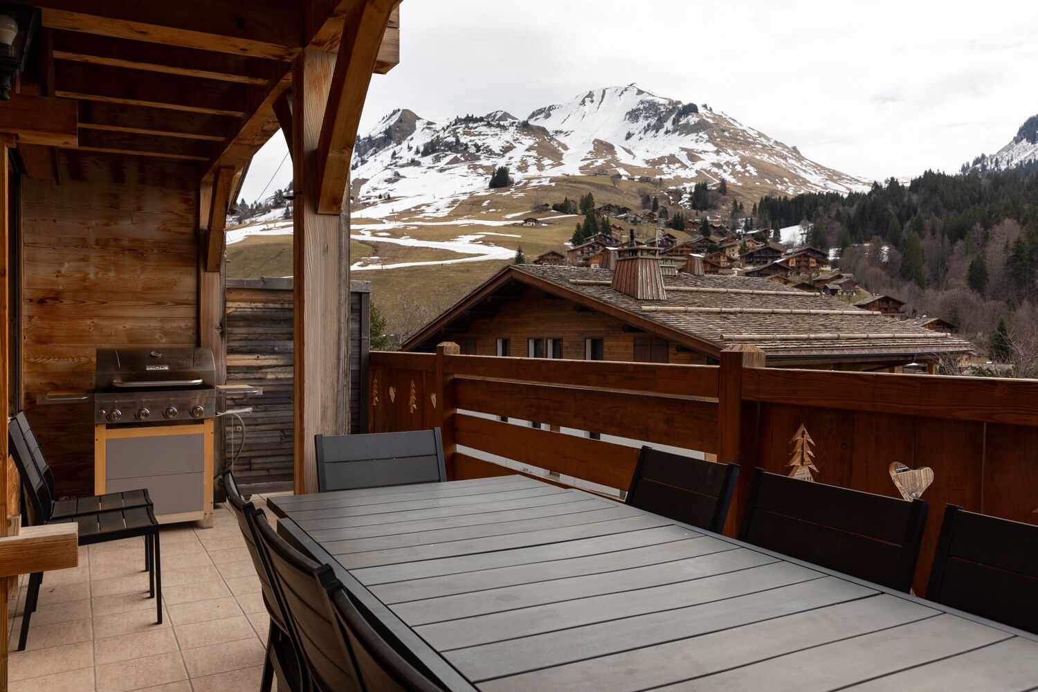 Une terrasse en bois avec un barbecue et un espace salon offrant une vue sur un village de montagne.