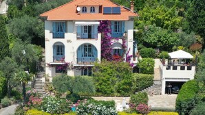 Une maison à deux étages, avec un toit recouvert de tuiles rouges, des murs blancs et des volets bleus. Elle possède un balcon surmonté d’un auvent blanc et est entourée d’une végétation luxuriante et de plantes en fleurs.