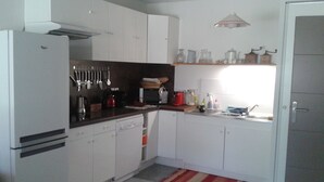 Una cocina compacta con armarios blancos, un refrigerador, un microondas y un lavamanos. Hay utensilios colgados en la pared, y un estante con frascos y una tetera.