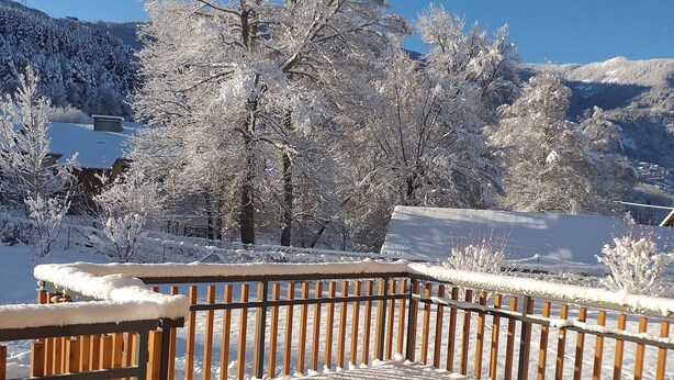 Una terraza cubierta de nieve con barandilla de madera, cielo azul despejado y árboles nevados al fondo.
