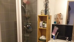 Un baño con ducha de vidrio, estante de madera con artículos de aseo y lavamanos blanco con espejo arriba.