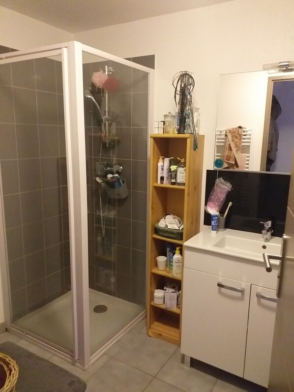 A bathroom with a glass-enclosed shower, a wooden shelf with toiletries, and a white sink with a mirror above it. - WEEKLY RENTALS, NIGHTS, summer-winter in GUILLESTRE (gateway to the Queyras) (Guillestre)