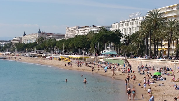 A beach with people sunbathing and swimming, palm trees, and buildings in the background. - Large 4P Pointe Croisette Huge terrace Sea view Beaches Wifi Air conditioning Garage (Cannes)