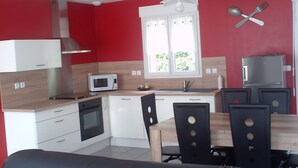 Une cuisine compacte avec des meubles blancs, un four à micro-ondes, une cuisinière et un évier. On y trouve une table à manger avec des chaises et une fenêtre avec des rideaux.