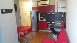 A compact kitchen with a refrigerator, microwave, and oven. There is a dining table with two chairs and a red tablecloth. The kitchen has red cabinets and a black countertop. - Rental Apartment F3 Fontenay-aux-Roses (Fontenay-aux-Roses)
