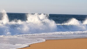 A sandy beach with footprints leading to the water, waves crashing, and a clear sky. - Villa d'ARCHITECTE, Sur les hauteurs, VUE MER, calme, TERRASSE 360°.   (casal mota)