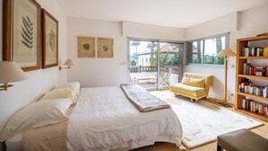 Un dormitorio con una cama, un sillón amarillo, una estantería y vistas al exterior.