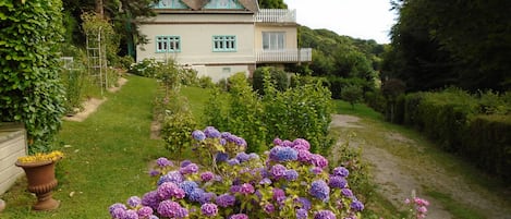 Una casa de fachada blanca y detalles verdes, rodeada de abundante vegetación y un jardín con flores moradas.