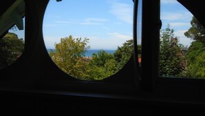 Vista a través de una ventana circular sobre un paisaje costero con árboles y el mar al fondo.
