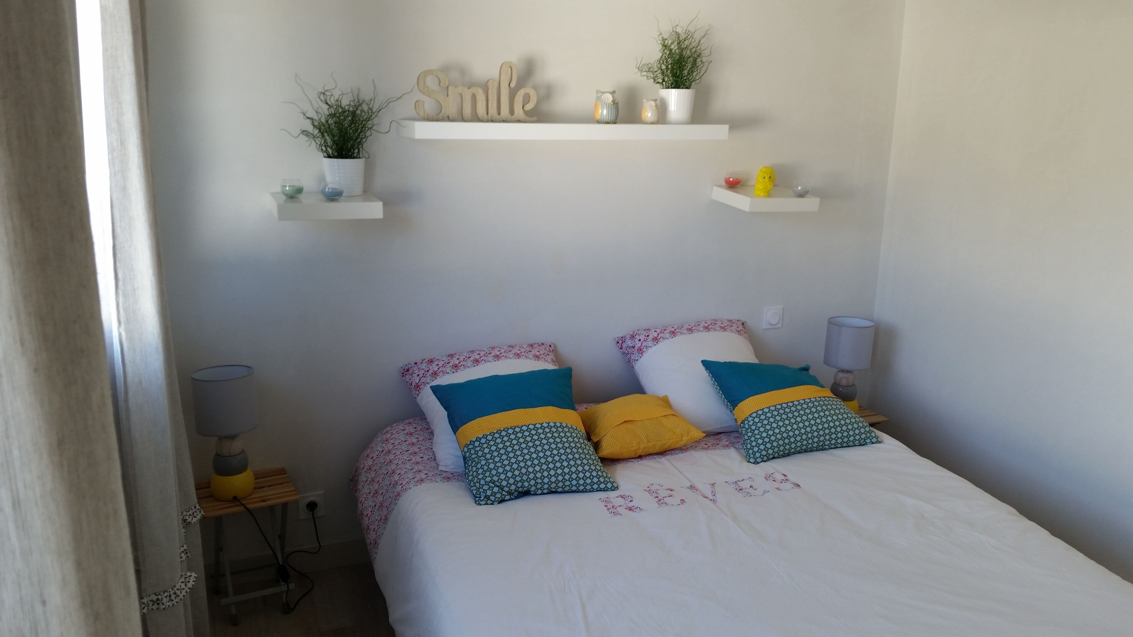 Un dormitorio con cama, almohadas, mesita de noche, lámpara y estantes en la pared con adornos.