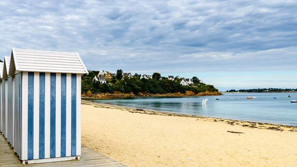 Ein Strand mit einer Reihe von blau-weiß gestreiften Strandhäusern, einem sandigen Ufer und einem ruhigen Meer mit Booten.