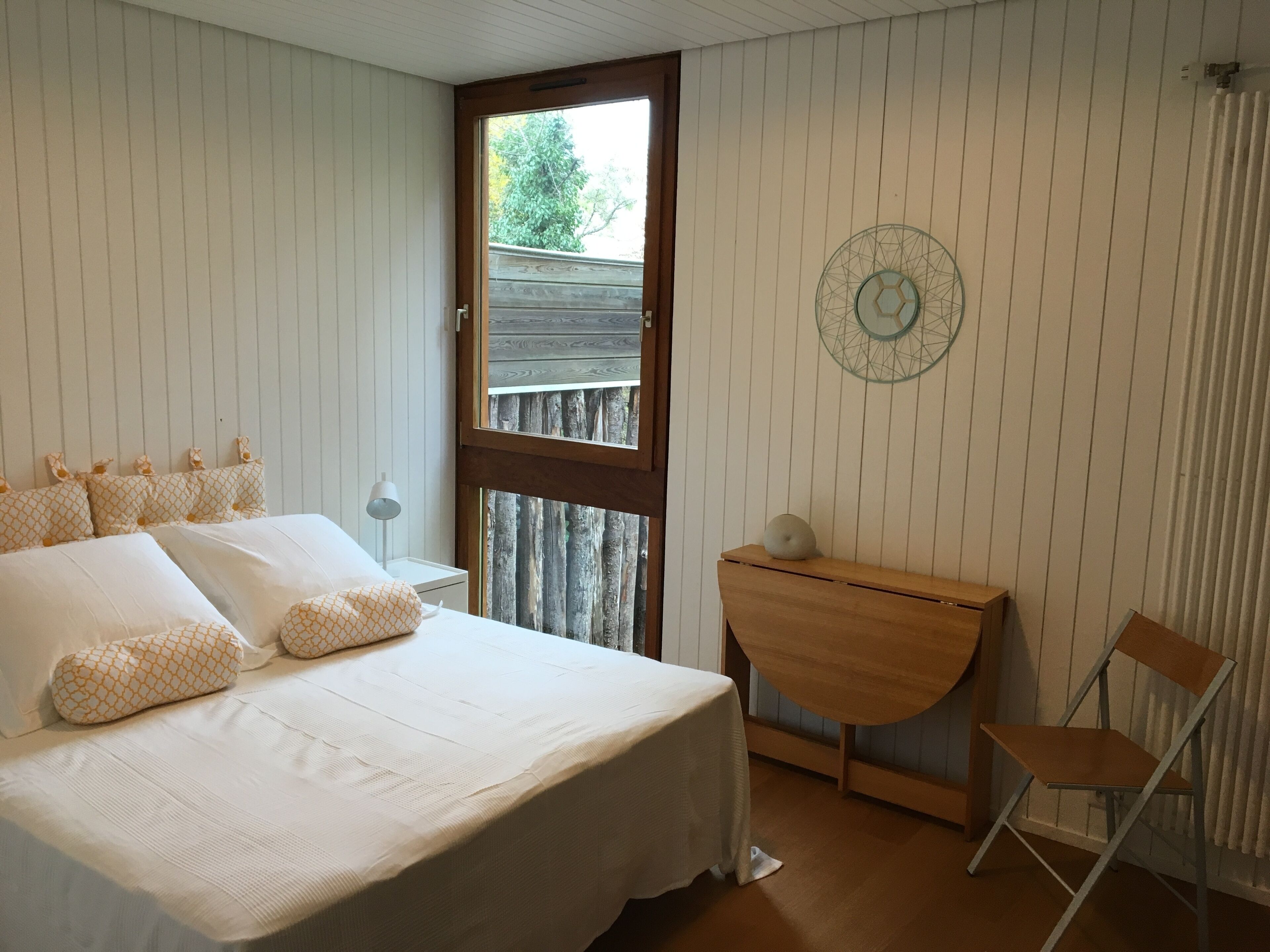 Une chambre à coucher avec un lit, un bureau en bois, une chaise, une fenêtre et un élément décoratif mural.