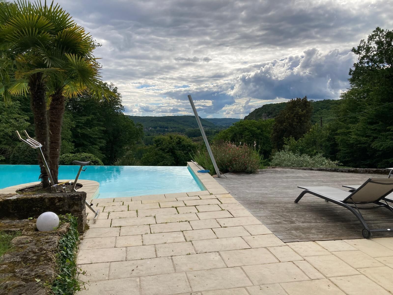 Ein Schwimmbecken mit Holzdeck und Liegestühlen, umgeben von Grünpflanzen und einer Bergkulisse.