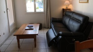 Una sala de estar con un sofá de cuero negro, una mesa de centro de madera, una lámpara y una ventana que ofrece vistas a zonas verdes y edificios.