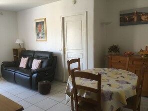Una sala de estar con un sofá de cuero negro, una mesa de comedor de madera con mantel floral y una puerta blanca.
