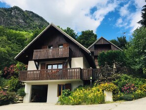 Un bâtiment de style chalet doté d’un balcon, entouré d’une végétation luxuriante et avec une chaîne de montagnes en toile de fond.