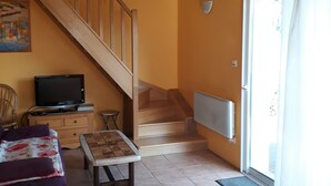 Een woonkamer met een houten trap, een televisie op een houten dressoir, een bank, een salontafel en uitzicht door een glazen deur.
