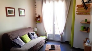 A cozy living room with a sofa, a coffee table, and a window with curtains. There are wall decorations and a clock. - Cozy vacation rental in the Arcachon Basin for 2–3 people: peaceful setting, amenities, and beaches (Gujan-Mestras)