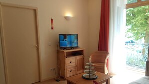 Une chambre d’hôtel avec un bureau en bois, une télévision, un fauteuil en osier et un balcon donnant sur la forêt.