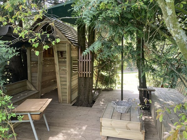 Una cabaña de madera con un banco y una mesa, rodeada de árboles y una terraza de madera.