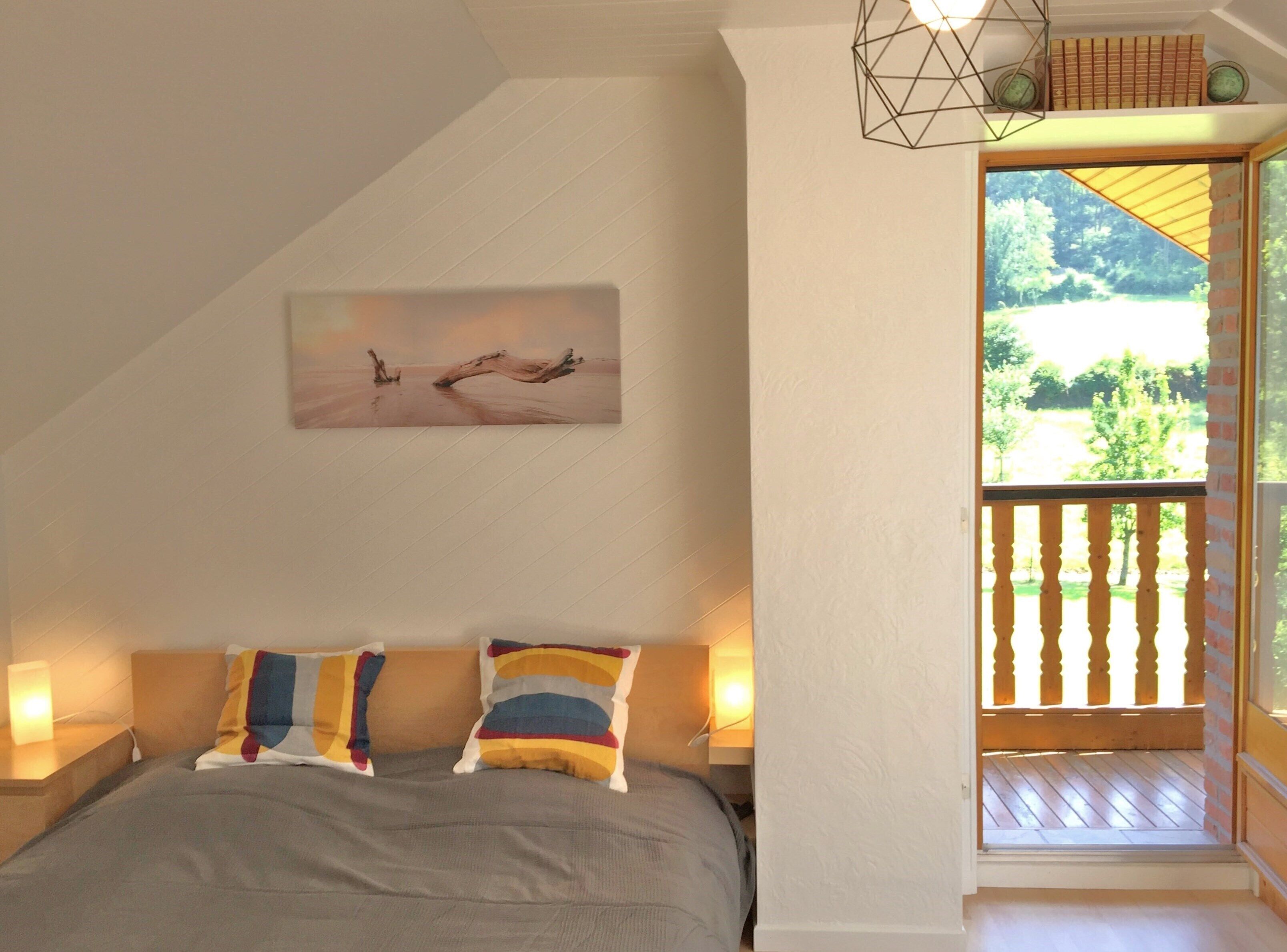 A bedroom with a bed, two pillows, a painting, a nightstand, a lamp, and a view of a balcony with a wooden railing.