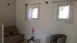 Un salon comprenant un canapé, un fauteuil, une petite table et un pot d’orchidée. Il y a deux fenêtres avec des rideaux.