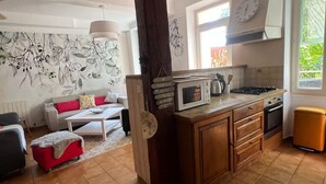 A cozy living room with a sofa, ottoman, and a coffee table. A kitchen area with a stove, microwave, and cabinets is visible. - Country house with large garden and swimming pool (Carces)