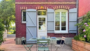 A wooden deck with a table set for dining, a brick building with shutters, and a garden with flowers. - Charming cottage with bay view and garden (Saint-Valery-Sur-Somme)