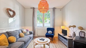A modern living room with a grey sofa, a blue armchair, a round wooden coffee table, and a TV stand with a lamp. - Charming cottage with bay view and garden (Saint-Valery-Sur-Somme)