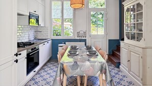 A kitchen with a dining table set for four, a hanging orange pendant light, and a view of a garden through the windows. - Charming cottage with bay view and garden (Saint-Valery-Sur-Somme)