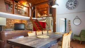 Een ruime woonkamer met een houten eettafel, comfortabele zitplaatsen, een stenen openhaard, een televisie en een trap naar een hoger gelegen verdieping.