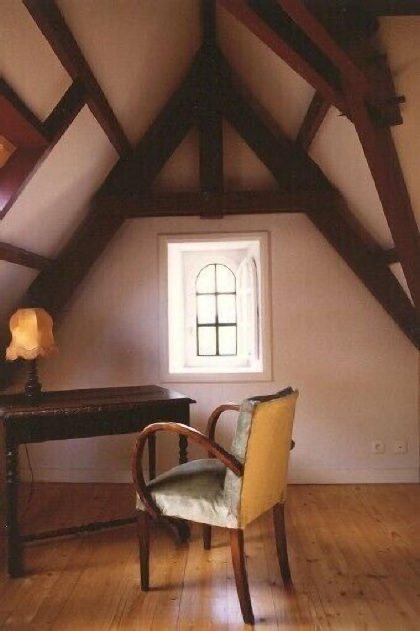 Una stanza con soffitto spiovente, una finestra con cornice bianca, una scrivania in legno e una sedia con cuscino.