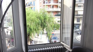 A room with two dark armchairs, a radiator, and a view of a street with buildings and parked cars.