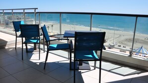 En balkon med blå stole og et bord, med udsigt over en strand og et klart hav med bygninger i det fjerne.