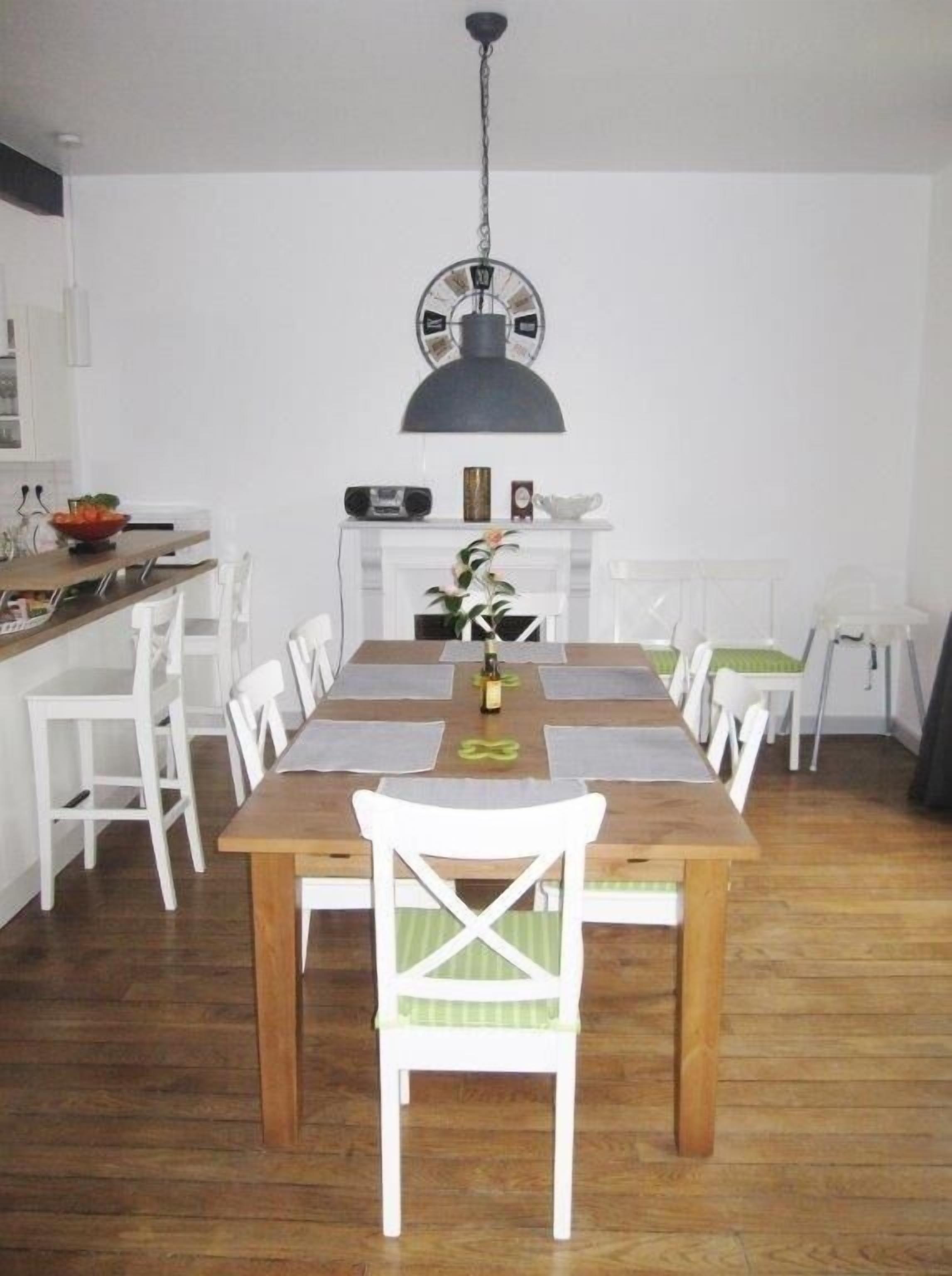 Un coin repas avec une table en bois et des chaises blanches, un luminaire suspendu et un meuble blanc contenant une radio et des objets décoratifs.