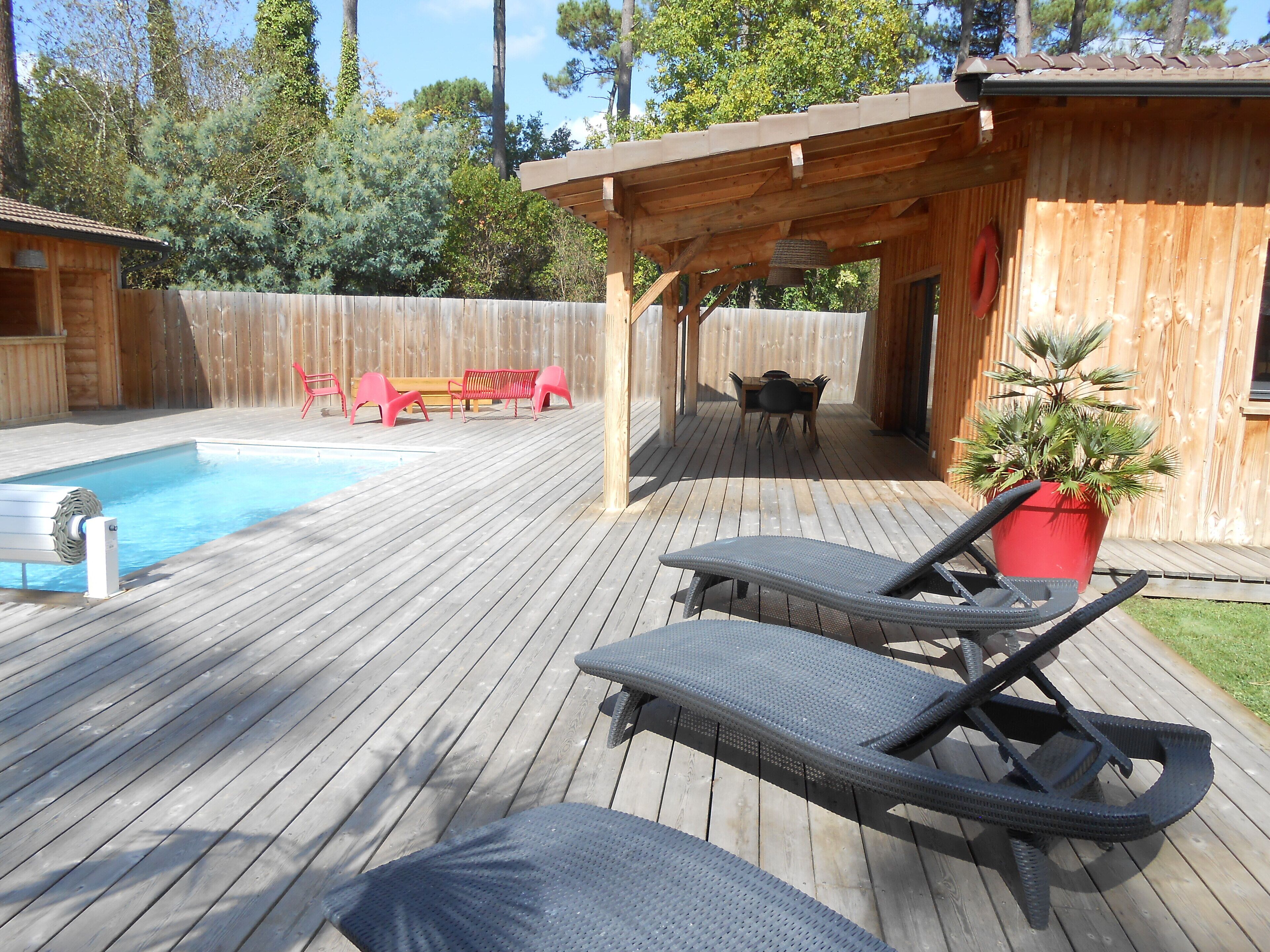 Um deck de madeira com piscina, espreguiçadeiras e área de estar coberta.