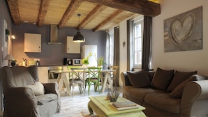 Een gezellige woonkamer met een bank, fauteuil en salontafel. Er is een eethoek met een tafel en stoelen, en een keuken op de achtergrond.