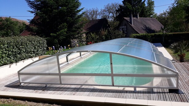 A covered swimming pool with a clear roof, surrounded by a wooden deck and lush greenery. - Old house located on an apple-red fruit farm (Savignac-Lédrier)