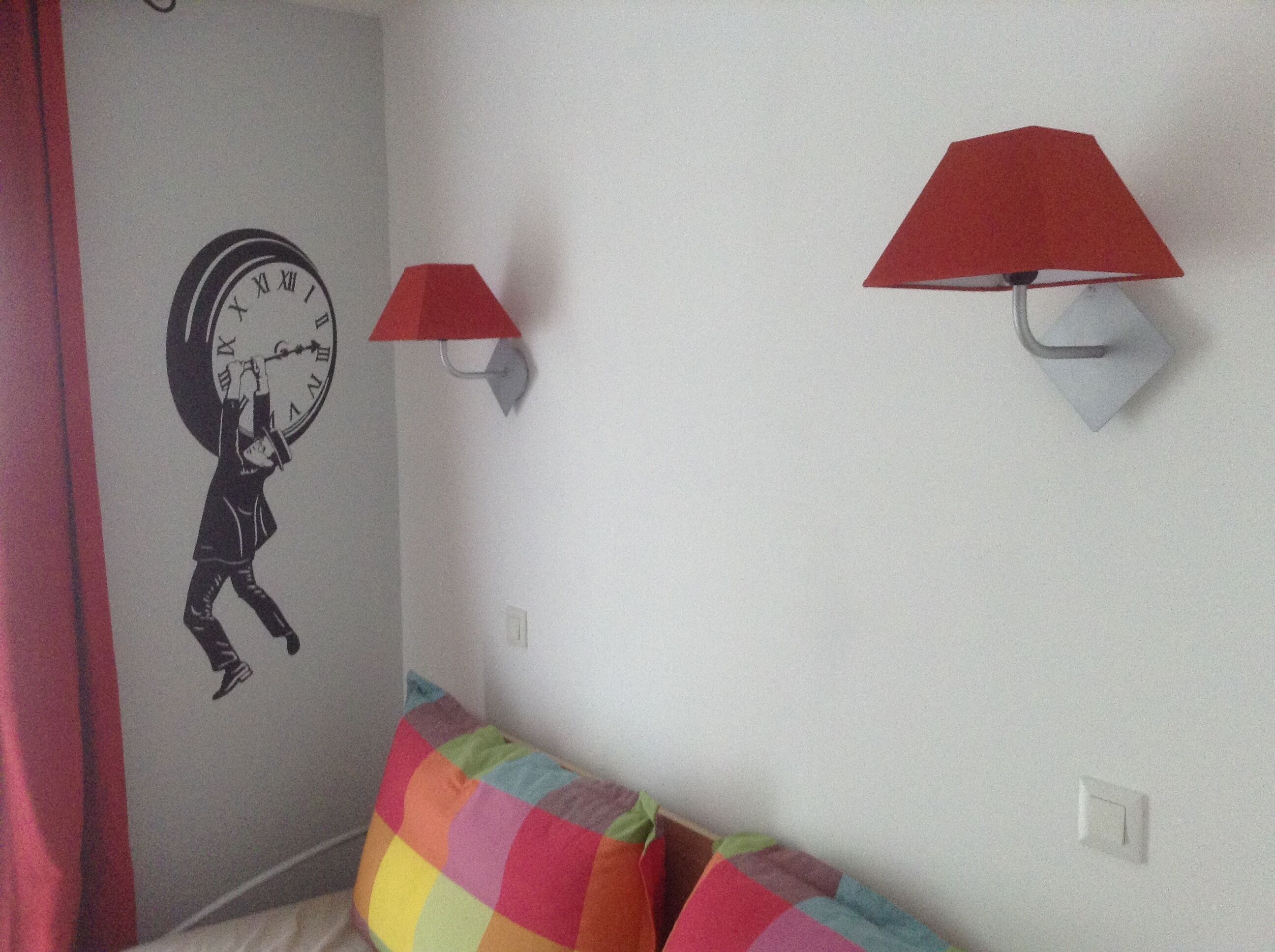 A room with a wall clock, two red lamps, and a colorful cushion on a sofa.