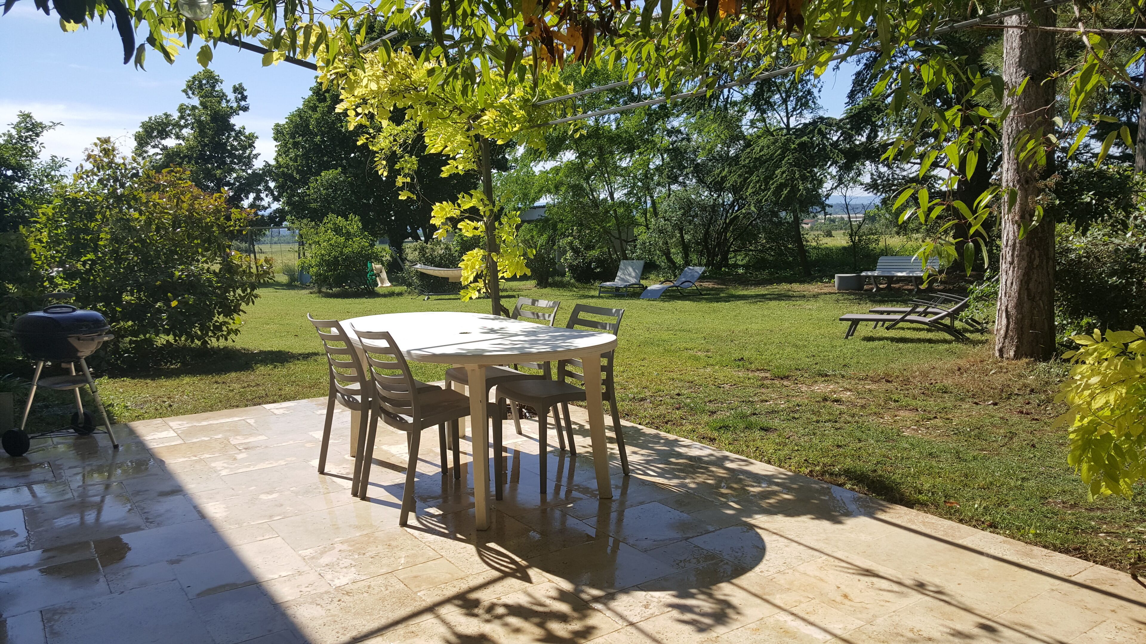 Une terrasse avec une table ronde et des chaises, entourée d’une pelouse verdoyante et d’arbres.