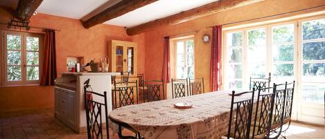 Un coin repas avec une table dressée pour six personnes, entourée de chaises dont le dossier est orné d’un motif en treillis. On y trouve également un coin cuisine avec un meuble blanc et une étagère suspendue au-dessus de l’évier.