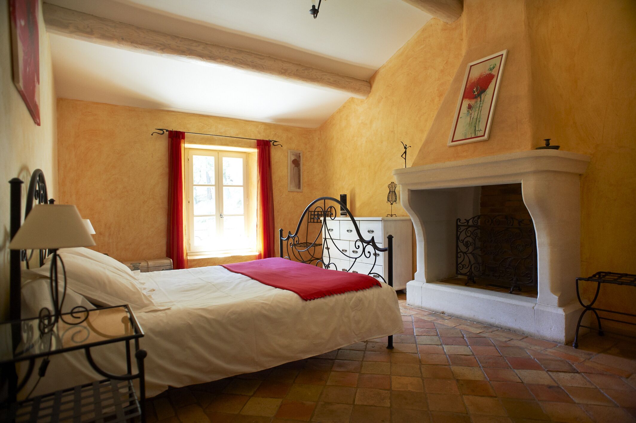 Un dormitorio con cama, mesita de noche, chimenea y ventana con cortinas rojas.