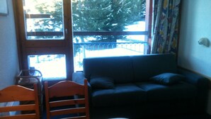 A room with a wooden table and chairs, a dark blue sofa, and a window with a view of trees and a railing. - Charming studio in Arcs 1800, at the foot of the slopes, in the heart of the village (Bourg-Saint-Maurice)