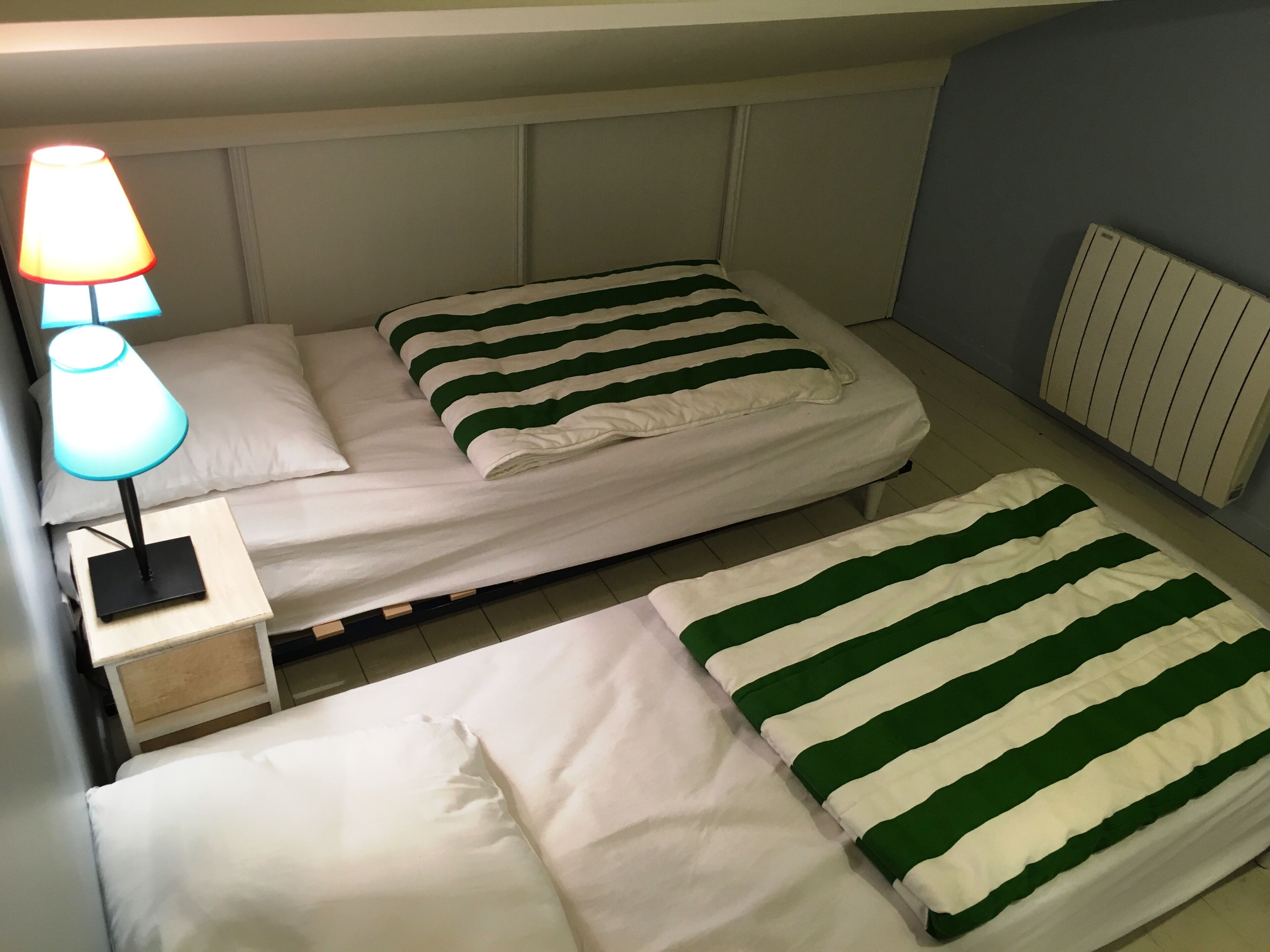 Et hotelværelse med to senge, hver med et grønt og hvidt stribet tæppe, et natbord med en lampe og en radiator på væggen.