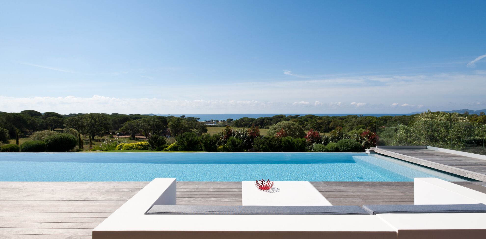 A swimming pool with a wooden deck and lounge chairs, surrounded by greenery and a view of the sea.