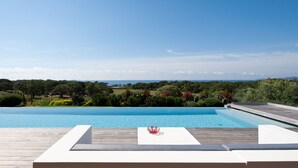 A swimming pool with a wooden deck and lounge chairs, surrounded by greenery and a view of the sea. - Magnificent villa with breathtaking views of Palombaggia beach Private sea access (Porto-Vecchio)