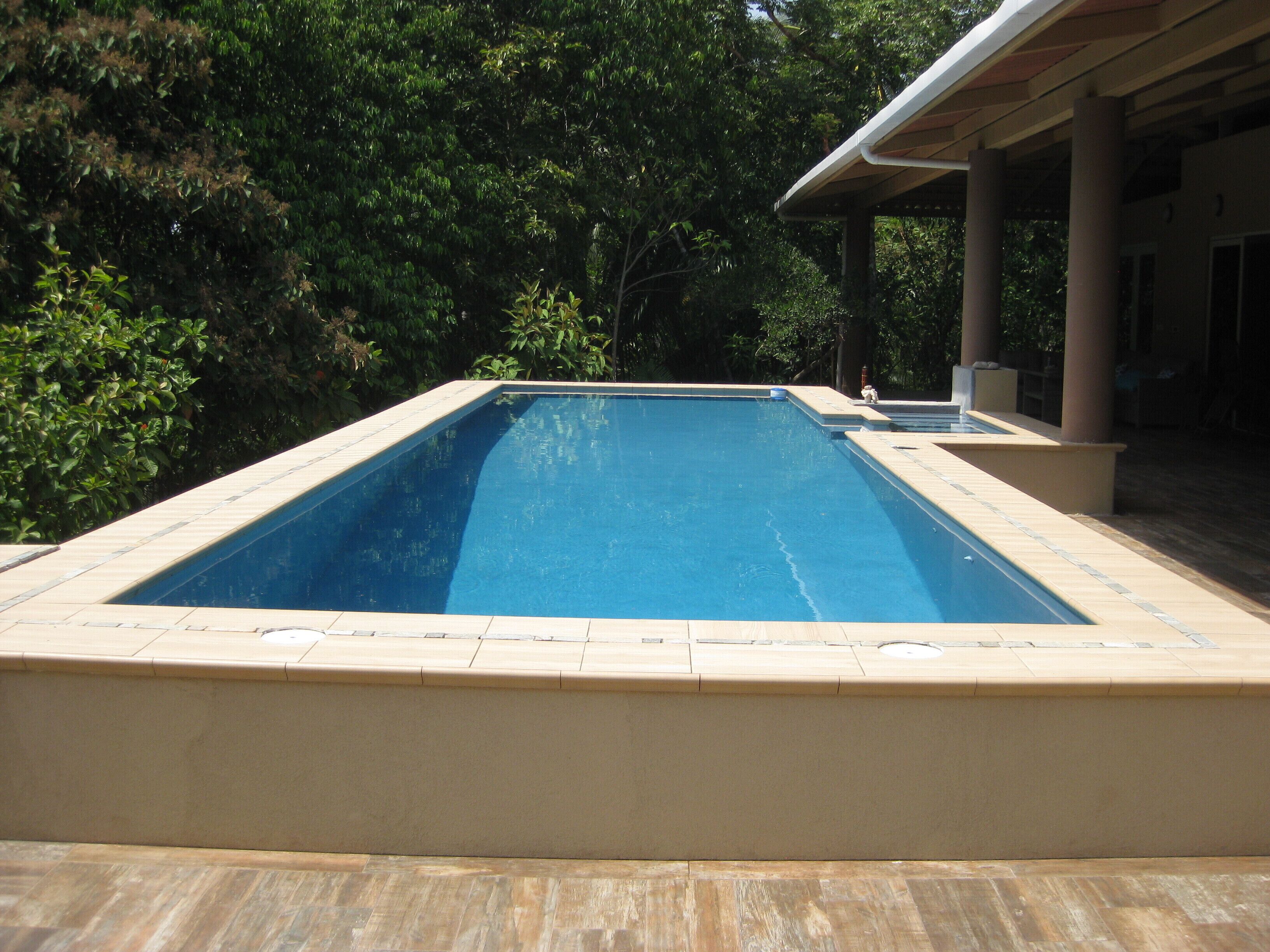 Una piscina rectangular con borde de azulejos, rodeada por una terraza de madera y un área de patio techada.